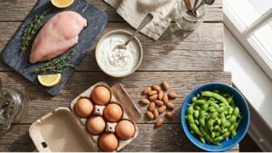 A rustic wooden table displaying various high-protein foods like eggs, Greek yogurt, chicken breast, and almonds.
